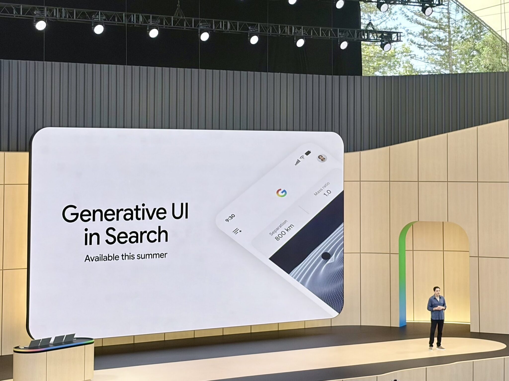 A presenter stands on stage at Google I/O next to a large screen displaying "Generative UI in Search Available this summer," featuring an image of a smartphone interface and highlighting advances in agentic search.