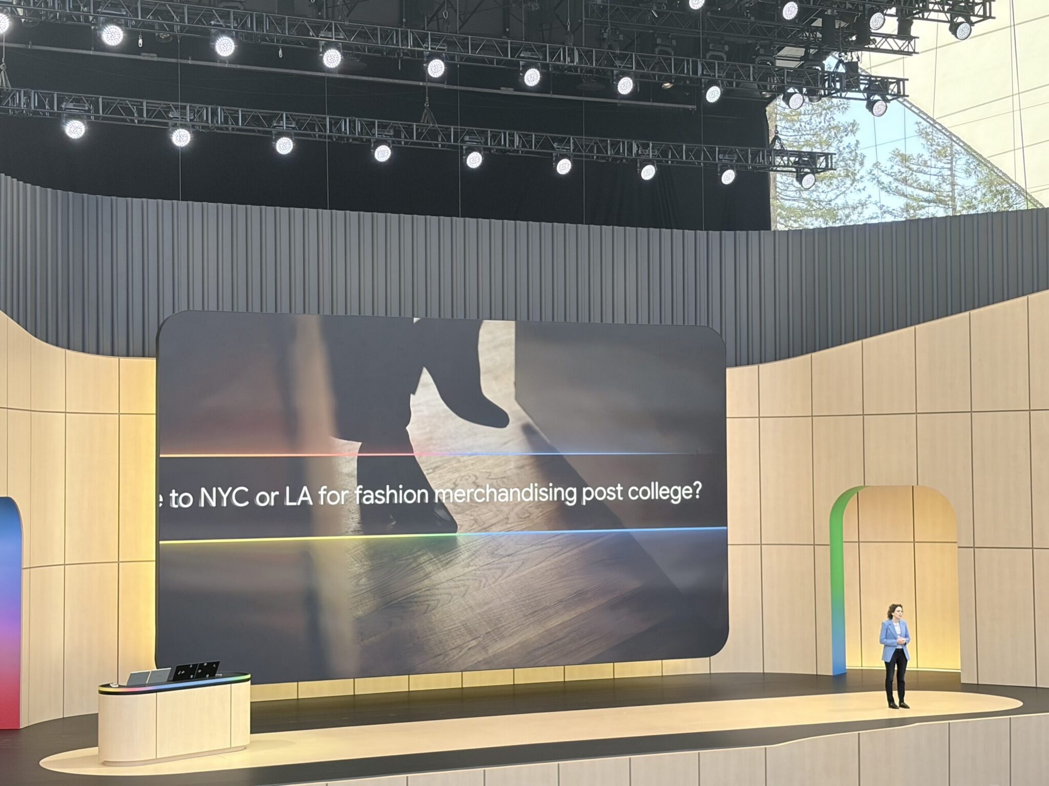 A person stands onstage at a modern indoor event, beside a large screen displaying text about choosing NYC or LA for fashion merchandising after college, reminiscent of presentations at Google I/O or talks on agentic search.
