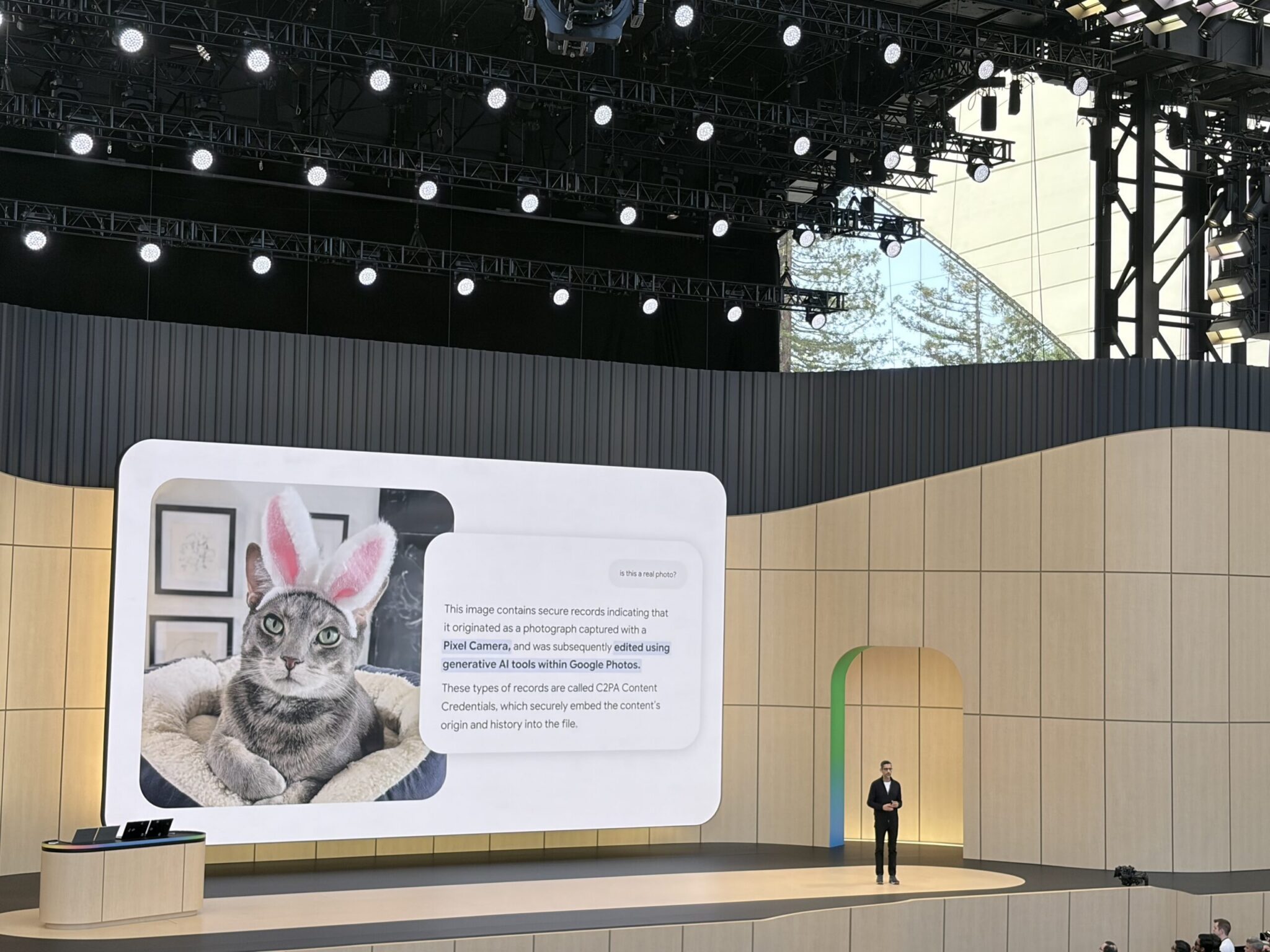 A presenter stands on stage at Google I/O beside a large screen displaying a cat in pink bunny ears, with a text box about AI-generated images, metadata, and the future of agentic search.