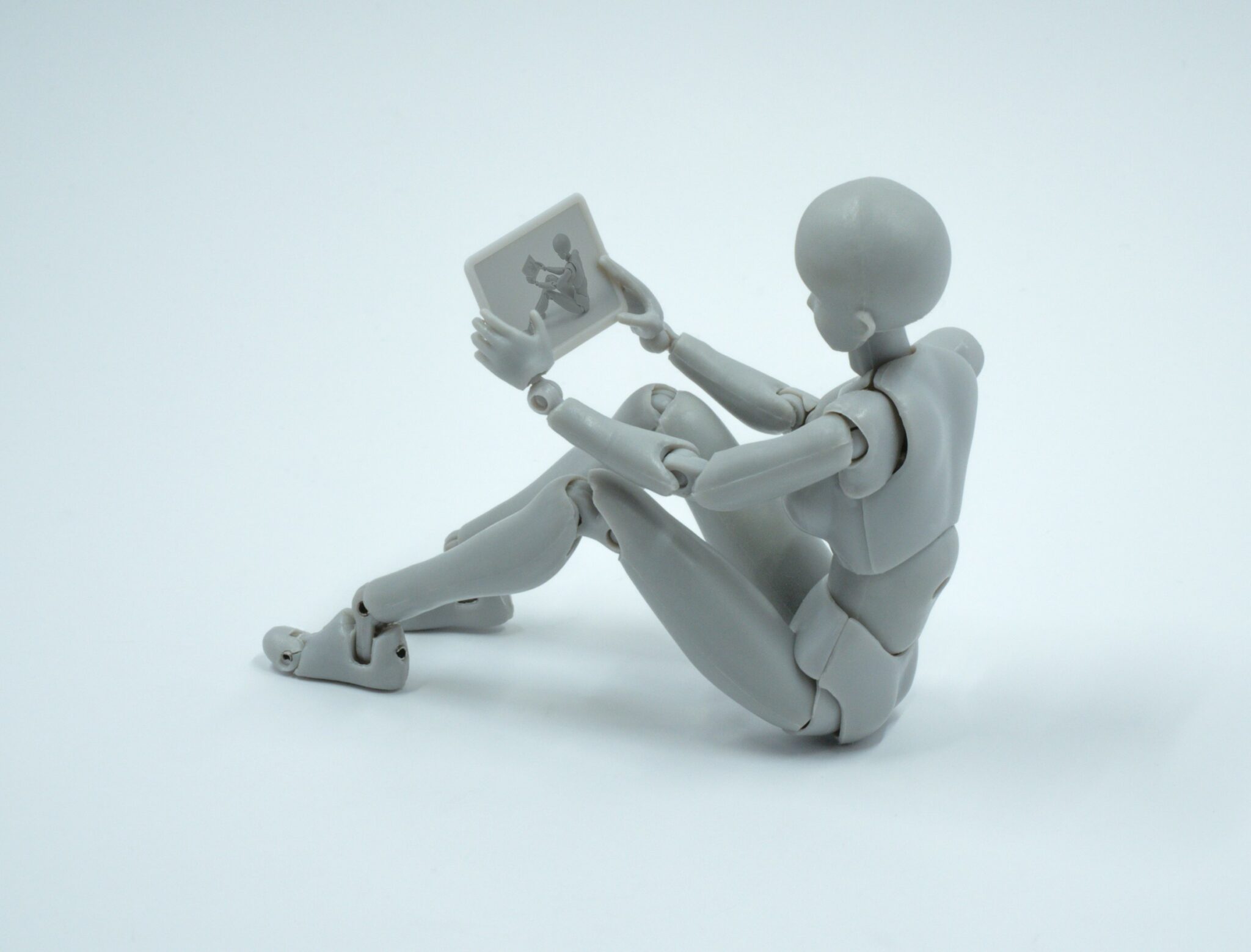 A gray humanoid figurine is sitting on the ground, holding and looking at a small rectangular tablet—perhaps exploring AI search optimization on its screen.