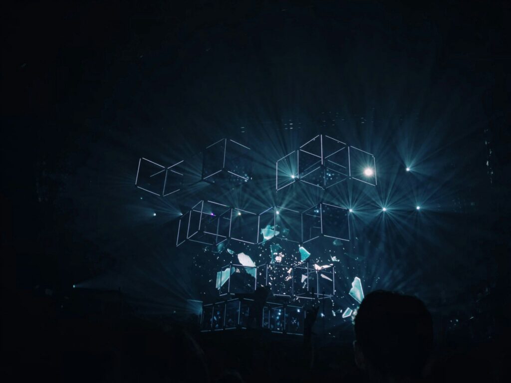 Stage setup featuring illuminated, floating cubes and blue lights, creating a captivating scene where you can build your own data experience, with silhouettes of audience members visible in the foreground.