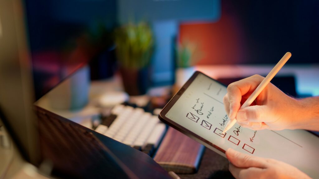 A person uses a stylus to check off items on a digital checklist displayed on a tablet, with a keyboard and monitor in the background—an important signal of organization and search efficiency.