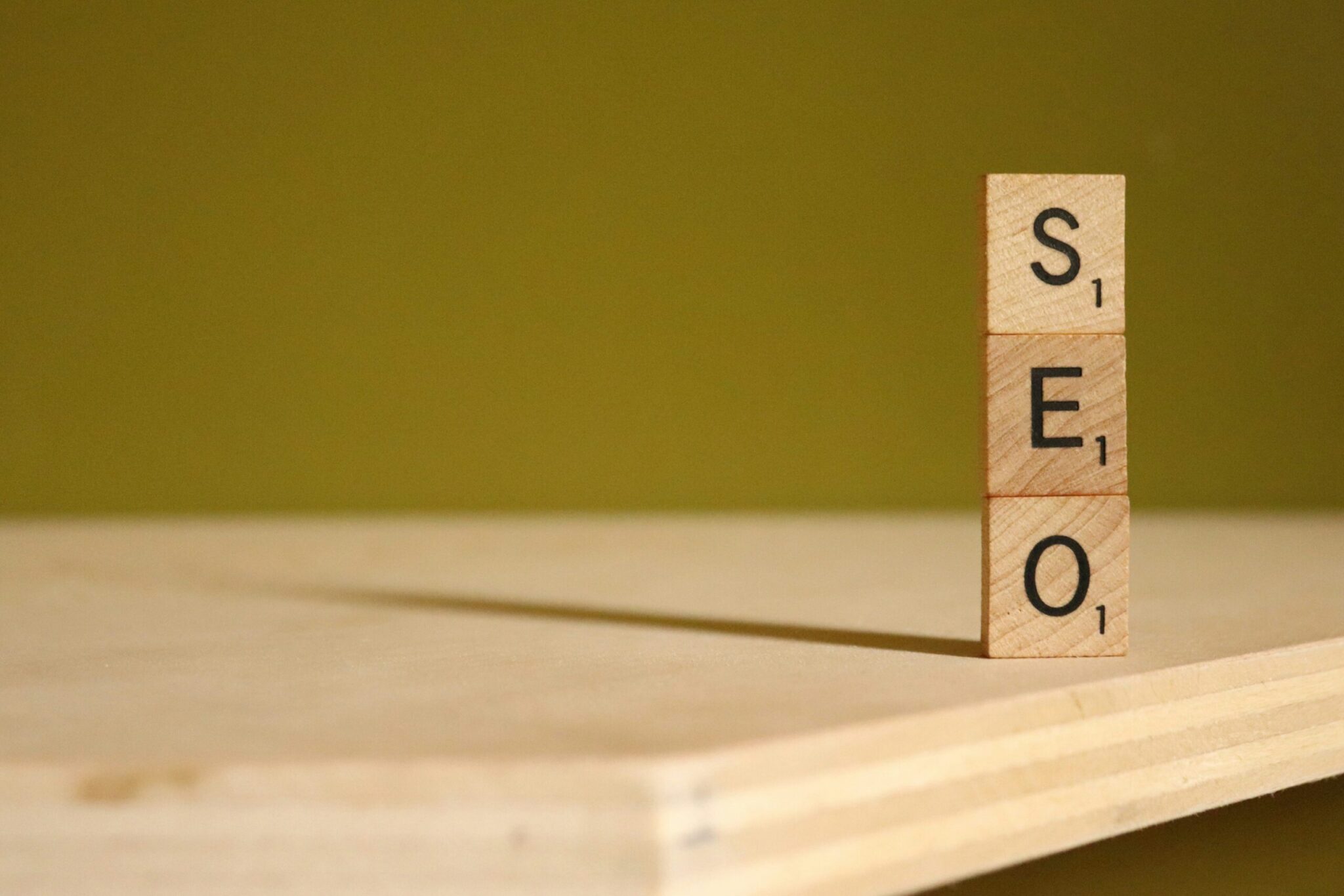 Three wooden Scrabble tiles stacked vertically on a wooden surface spell "SEO" against a plain green background, highlighting SEO fundamentals essential for success in search.
