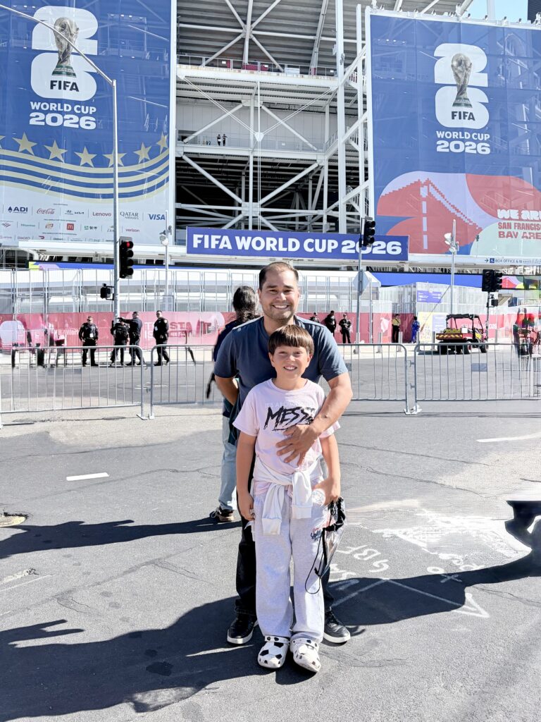 A man and a boy smile in front of FIFA World Cup 2026 banners outside a stadium on a sunny day.