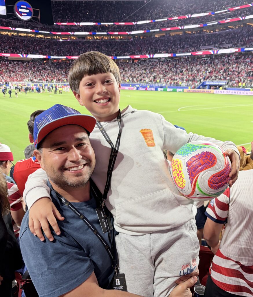 Smiling man and boy at a soccer stadium, boy holding a colorful soccer ball, crowd in the background.