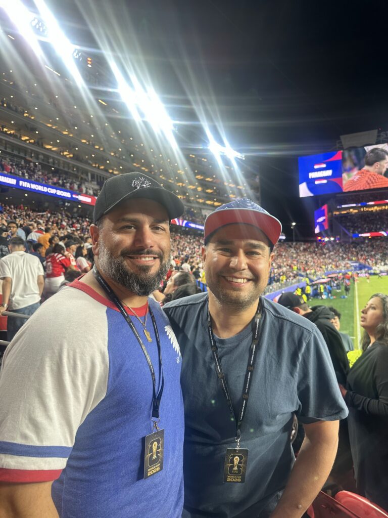 Two smiling men wearing lanyards at a crowded stadium during a nighttime FIFA World Cup event, capturing the excitement reflected in world cup search trends.