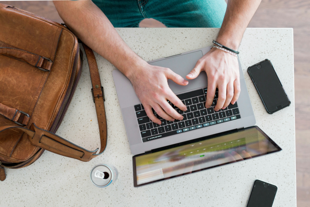 Person typing on a laptop at a table with a bag, phone, tablet, and a can of drink.