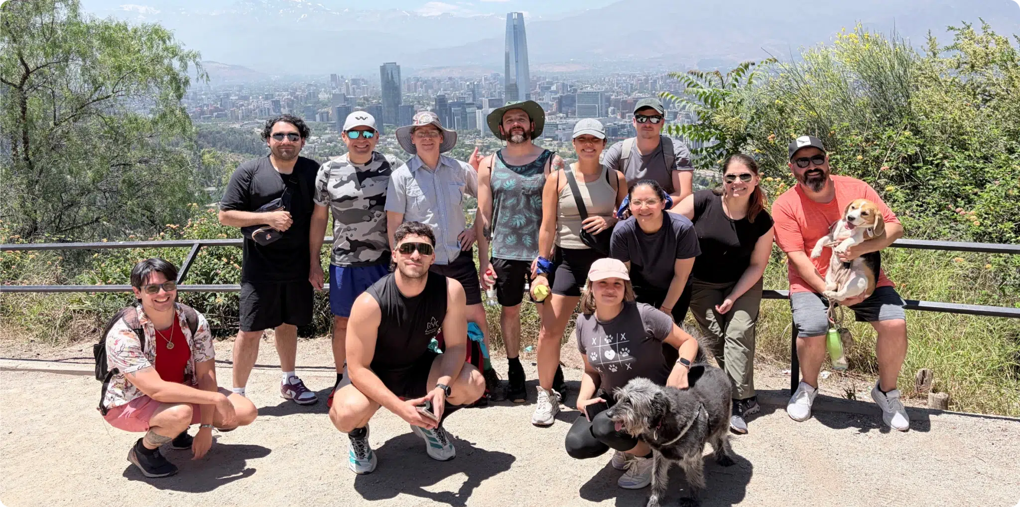 Group of people and two dogs posing outdoors with a city skyline and mountains in the background.