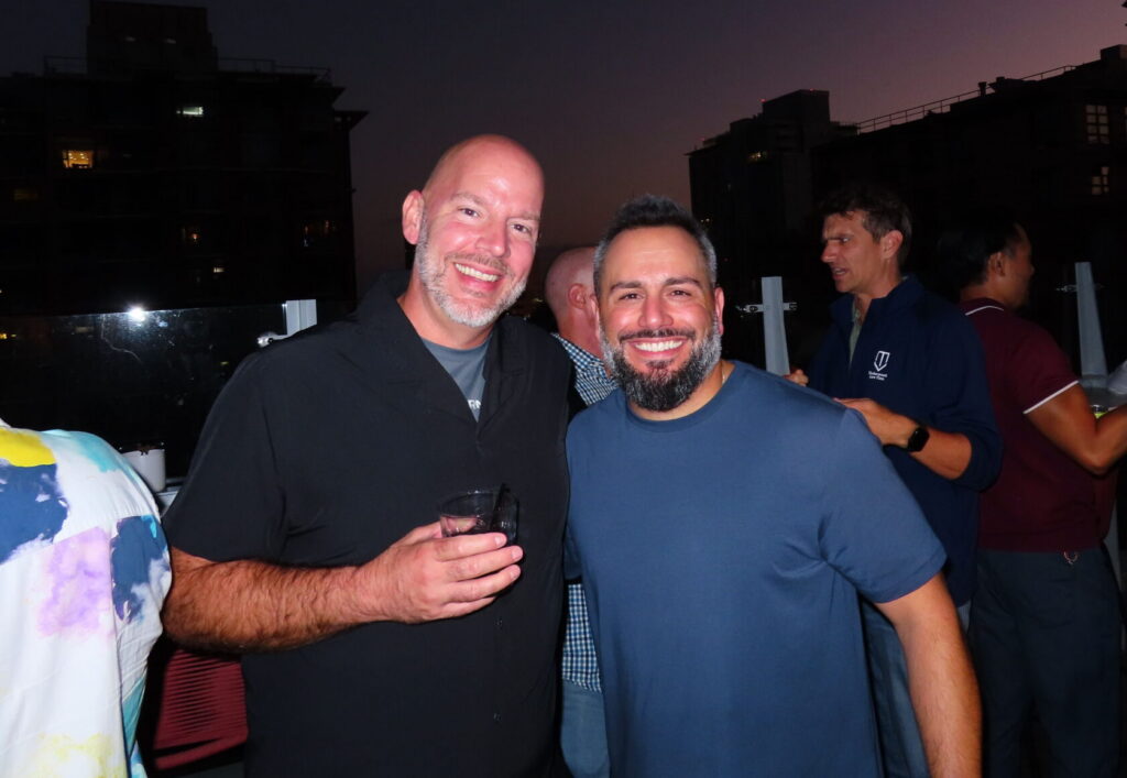Two smiling men pose together at an outdoor evening gathering with city buildings in the background.