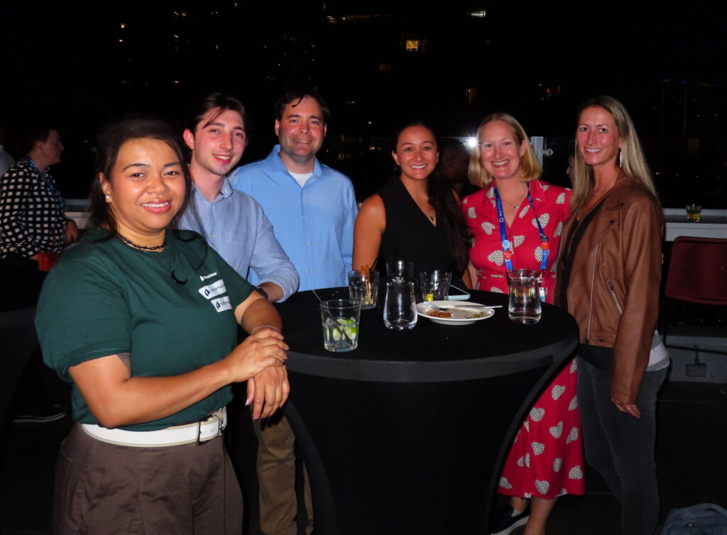 Six people smile around a tall black table with drinks at an outdoor nighttime event.