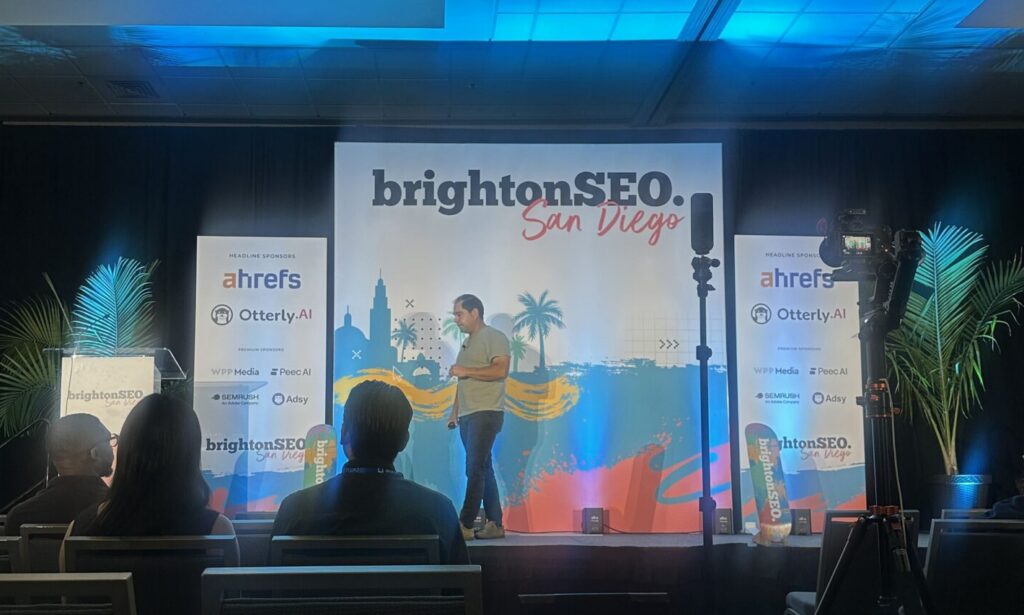 A speaker presents on stage at brightonSEO San Diego, with banners and an audience in a conference room.