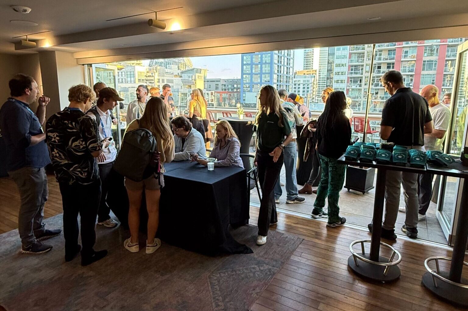 People gather around a table near large windows overlooking a cityscape in a modern indoor space.