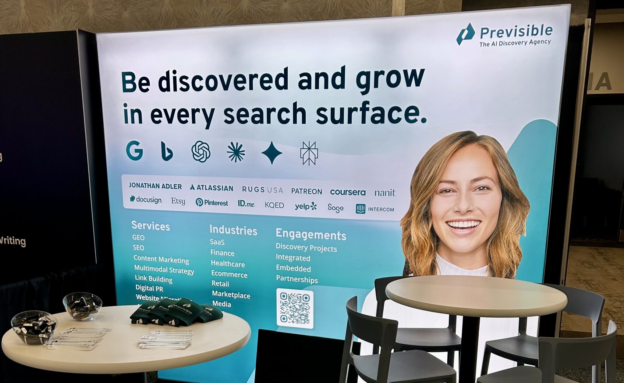 Trade show booth with a smiling woman, company logos, and text about AI-driven discovery services and industries; the display also features a BrightonSEO recap highlighting recent innovations in search marketing.