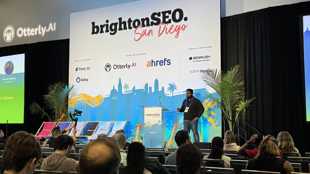 A speaker presents at brightonSEO San Diego; audience listens, with sponsor logos and palm plants on stage.